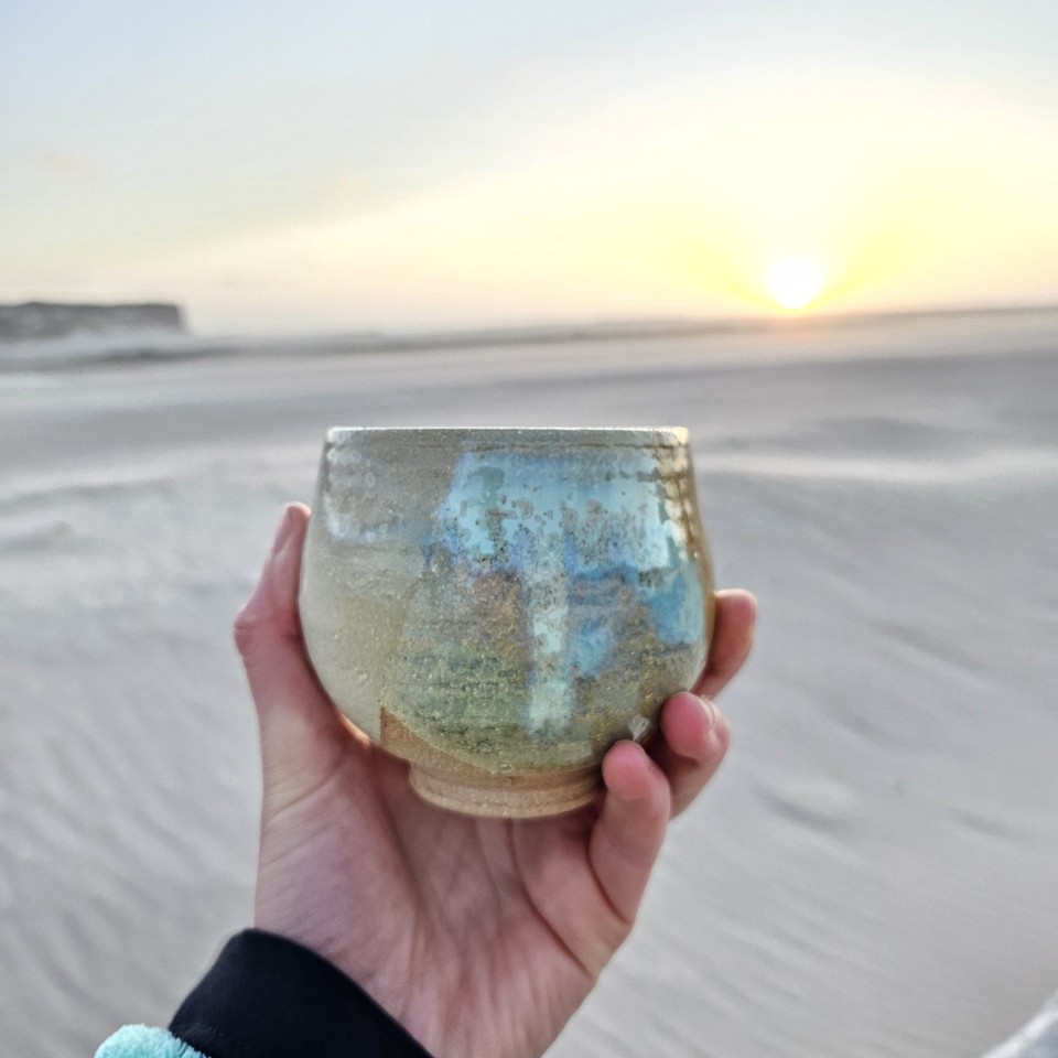 A handmade glazed cup held up on the beach at sunset
